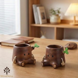 Puede incluir: Dos macetas de madera con forma de tocón de árbol, con caras de dibujos animados y detalles de hojas verdes. Las macetas son marrones con un acabado liso y tienen pequeños pies redondeados. Las macetas están sobre una mesa de madera.