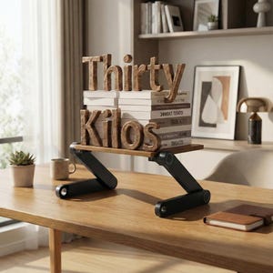 Puede incluir: Un organizador de escritorio de madera con las palabras "Thirty Kilos" encima de una pila de libros. El organizador tiene una base negra ajustable y est&aacute; sobre una mesa de madera. Una peque&ntilde;a planta suculenta y una taza de caf&eacute; tambi&eacute;n est&aacute;n sobre la mesa.