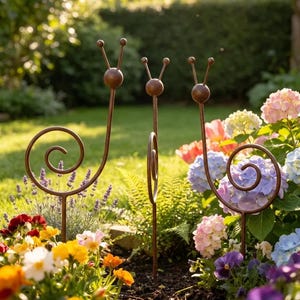 May include: Three brown metal garden stakes shaped like snails with spiral shells and long antennae. Each stake features a thin vertical rod base, a spherical head with two protruding stalks, and a circular wire coil representing the snail shell.
