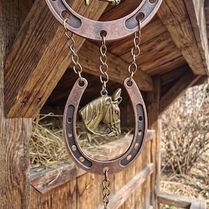 May include: A decorative hanging ornament featuring two copper-toned metal horseshoes and two brass-colored horse head reliefs. The pieces are connected by metal chains, with a bell suspended at the bottom. The finish is weathered and metallic.