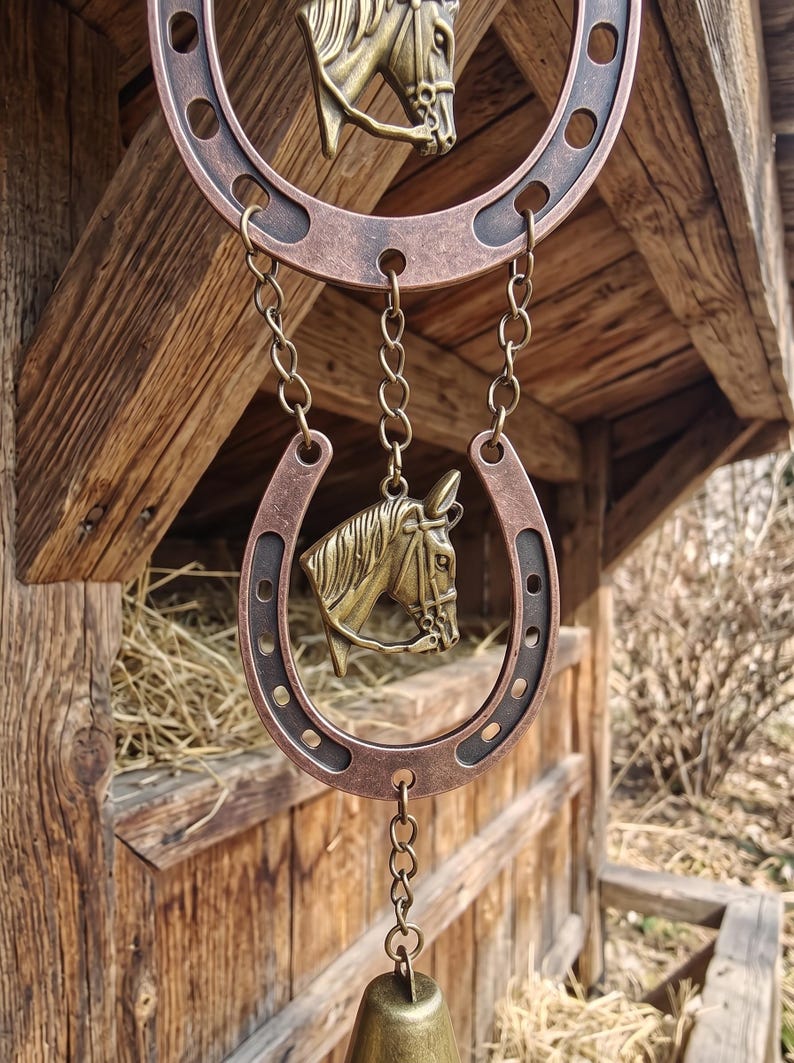 May include: A decorative hanging ornament featuring two copper-toned metal horseshoes and two brass-colored horse head reliefs. The pieces are connected by metal chains, with a bell suspended at the bottom. The finish is weathered and metallic.