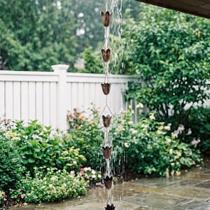 May include: A copper rain chain featuring a series of interconnected tulip-shaped cups suspended from a roof gutter. Water flows down through the metal cups, creating a vertical stream that lands on a large, smooth, rounded stone base.