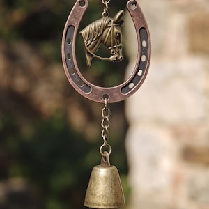 May include: A hanging ornament featuring a copper-toned horseshoe with a bronze-colored horse head emblem centered inside. A metal chain connects the horseshoe to a small, weathered brass bell at the bottom, creating a vertical decorative piece.