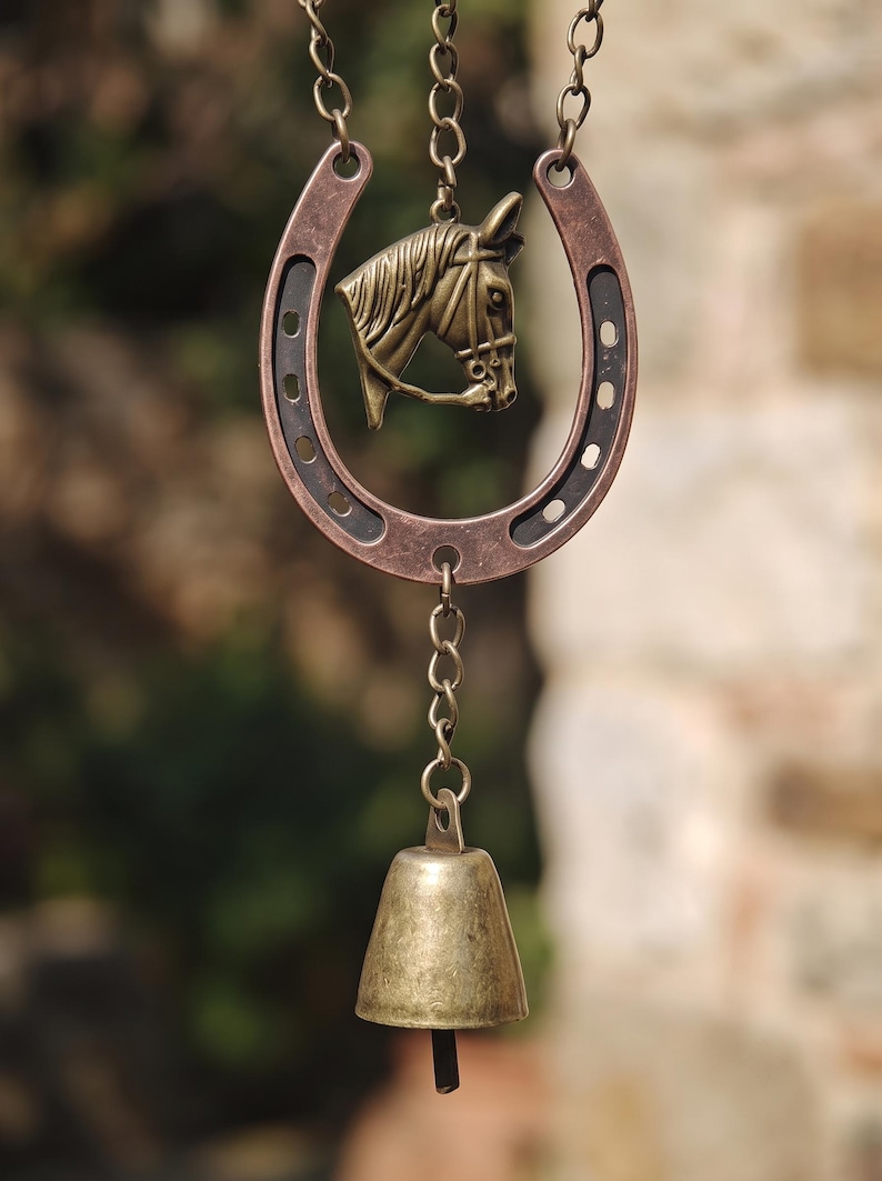 May include: A hanging ornament featuring a copper-toned horseshoe with a bronze-colored horse head emblem centered inside. A metal chain connects the horseshoe to a small, weathered brass bell at the bottom, creating a vertical decorative piece.