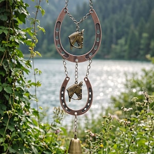 May include: A hanging ornament featuring two copper-toned metal horseshoes connected by chains. Each horseshoe contains a brass-colored horse head charm in the center. A small brass bell hangs from the bottom of the lower horseshoe.