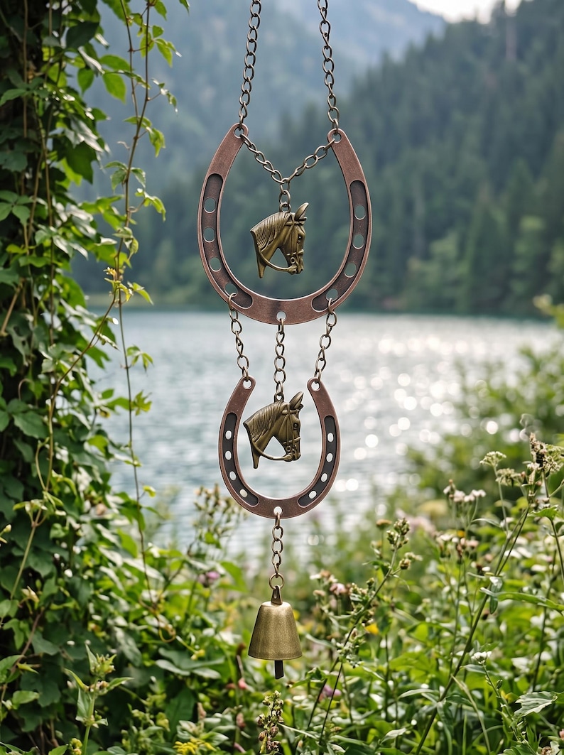 May include: A hanging ornament featuring two copper-toned metal horseshoes connected by chains. Each horseshoe contains a brass-colored horse head charm in the center. A small brass bell hangs from the bottom of the lower horseshoe.