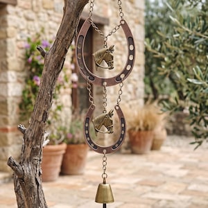 May include: A hanging decorative ornament featuring two copper-toned metal horseshoes connected by chains. Each horseshoe contains a brass-colored horse head emblem in the center. A small brass bell hangs from the bottom of the lower horseshoe.