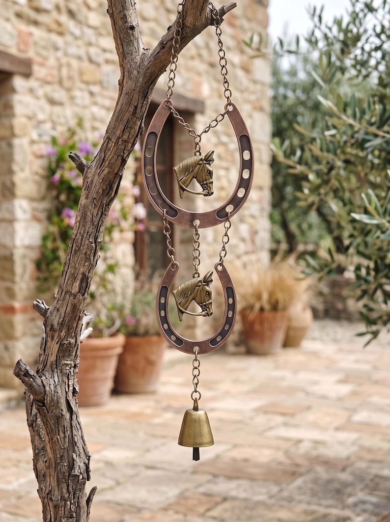 May include: A hanging decorative ornament featuring two copper-toned metal horseshoes connected by chains. Each horseshoe contains a brass-colored horse head emblem in the center. A small brass bell hangs from the bottom of the lower horseshoe.