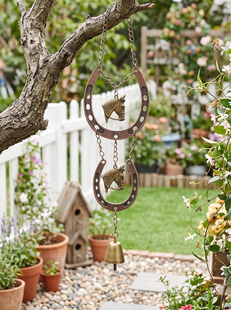 May include: A hanging wind chime featuring two copper-toned metal horseshoes arranged vertically, each centered with a bronze-colored horse head relief. The pieces are connected by metal chains, with a small brass bell suspended at the bottom.