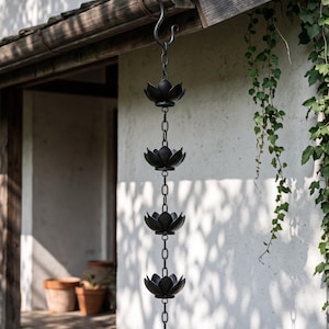 Op de afbeelding: Een zwarte metalen regenketting met een reeks lotusbloemvormige bekers verbonden door verticale schakels. De ketting hangt aan een gebogen metalen haak die is bevestigd aan de rand van een houten dakoverstek en hangt verticaal naar beneden.