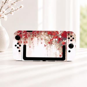 May include: A handheld gaming console featuring a decorative skin with a watercolor-style print of red and white roses, green vines, and small butterflies. The device has white controllers on each side with black buttons and joysticks.