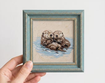 May include: A square oil painting featuring two brown sea otters floating in blue water, enclosed in a textured, distressed light blue wooden frame with gold inner detailing. The otters are depicted with closed eyes, holding paws together.