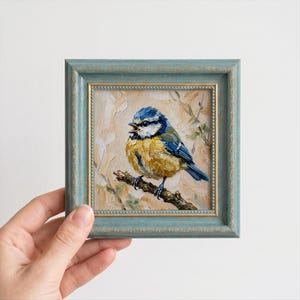 Pode incluir: Uma pintura a óleo de um chapim-azul pousado num ramo, feita com pinceladas visíveis. O pássaro apresenta uma coroa azul, peito amarelo e bochechas brancas. Está inserido numa moldura decorativa azul-clara com um rebordo dourado.