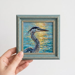 May include: A small framed oil painting of a grey heron with a long neck and sharp beak. The bird is set against a background of shimmering blue water reflecting golden sunlight. The artwork is encased in a textured blue wooden frame.