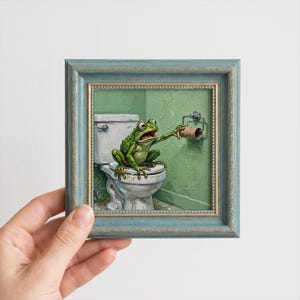 May include: A framed oil painting depicting a green frog sitting on a white toilet in a green-walled bathroom. The frog is reaching out with one hand towards an empty brown cardboard toilet paper roll holder mounted on the wall beside the toilet.