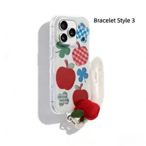 May include: Clear phone case with a white, apple-themed design. The case features red apples, blue and green floral accents, and the text "Bracelet Style 3". A fluffy white wrist strap and a felt apple charm are attached.