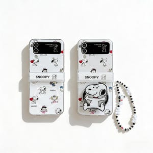 May include: Two white phone cases with a Snoopy design. The cases feature Snoopy and other Peanuts characters, hearts, and the word "Snoopy". One case has a Snoopy charm and a black and white beaded wrist strap.