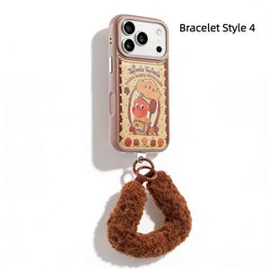 May include: A rose-gold phone case with a cartoon graphic and a brown wrist strap. The case features a whimsical illustration of a chef character. The wrist strap is made of a soft, fuzzy material.