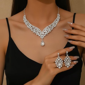 Op de afbeelding: Een zilverkleurige sieradenset met een ketting en oorbellen. De ketting heeft een V-vormig ontwerp met talrijke heldere, sprankelende stenen en een druppelvormige hanger. De oorbellen weerspiegelen de vorm van de hanger.
