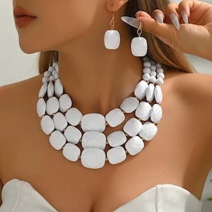 May include: A white statement necklace and matching earrings set. The necklace features three strands of large, geometric, white beads. The earrings have a similar design, with a single white geometric bead suspended from a gold-colored hook.