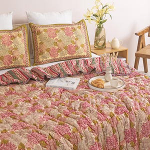 Puede incluir: Una cama con un edredón con estampado floral y cojines a juego. El edredón y los cojines presentan un diseño floral rosa, verde y crema. Una bandeja con un cruasán y una taza de café descansa sobre la cama. Un jarrón con flores amarillas está sobre una mesita de noche.