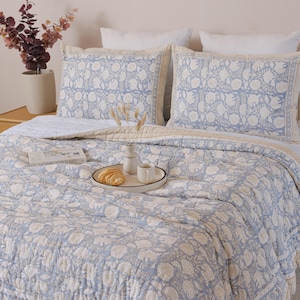 May include: A bed with a light blue floral patterned quilt and matching pillowcases. A tray with a croissant and coffee cup sits on the quilt. A vase of flowers and a book are on the nightstand.