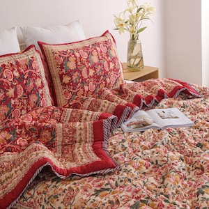 May include: A quilted bedspread and two matching pillow shams featuring a vibrant red, cream, and green floral block print pattern. The bedding has a distinct border design with a thin striped edge, displayed on a bed with an open book.
