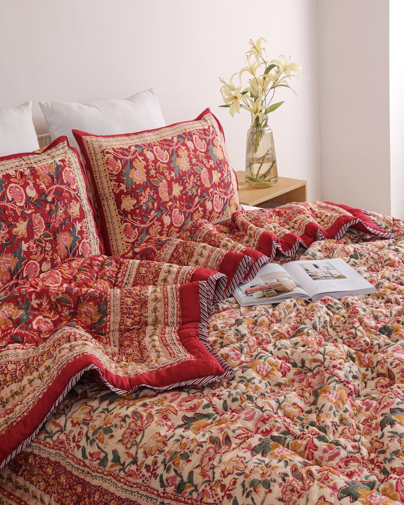 May include: A quilted bedspread and two matching pillow shams featuring a vibrant red, cream, and green floral block print pattern. The bedding has a distinct border design with a thin striped edge, displayed on a bed with an open book.
