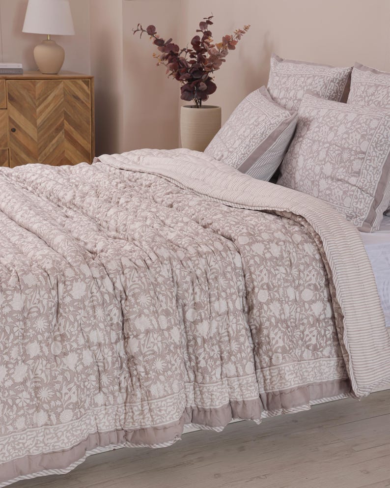 May include: A beige and white floral patterned quilt and matching pillow set on a bed. The quilt features a detailed floral design with a border. The pillows have a similar pattern and a striped border. The bedding set is a neutral color.
