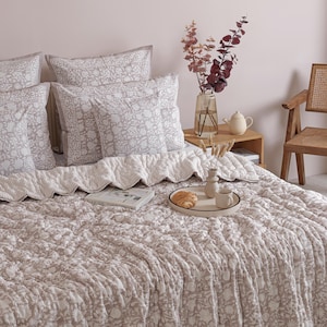May include: A bed with a floral patterned comforter and matching pillows in shades of beige and white. A tray with a croissant and coffee cup sits on the bed. A vase of flowers and a small table are also visible.
