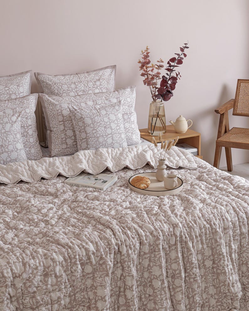 May include: A bed with a floral patterned comforter and matching pillows in shades of beige and white. A tray with a croissant and coffee cup sits on the bed. A vase of flowers and a small table are also visible.