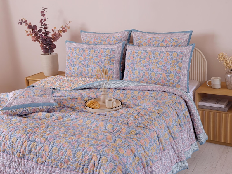 May include: A bed is made with a floral patterned quilt and matching pillowcases in shades of blue, peach, and yellow. A tray with a croissant and coffee cups sits on the quilt. The bed is in a bedroom with a neutral color palette.