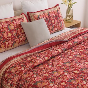 May include: A red quilted bedspread and two matching rectangular pillow shams featuring a dense floral and pomegranate pattern in shades of pink, orange, and green. A solid light gray square throw pillow sits between the two patterned shams.