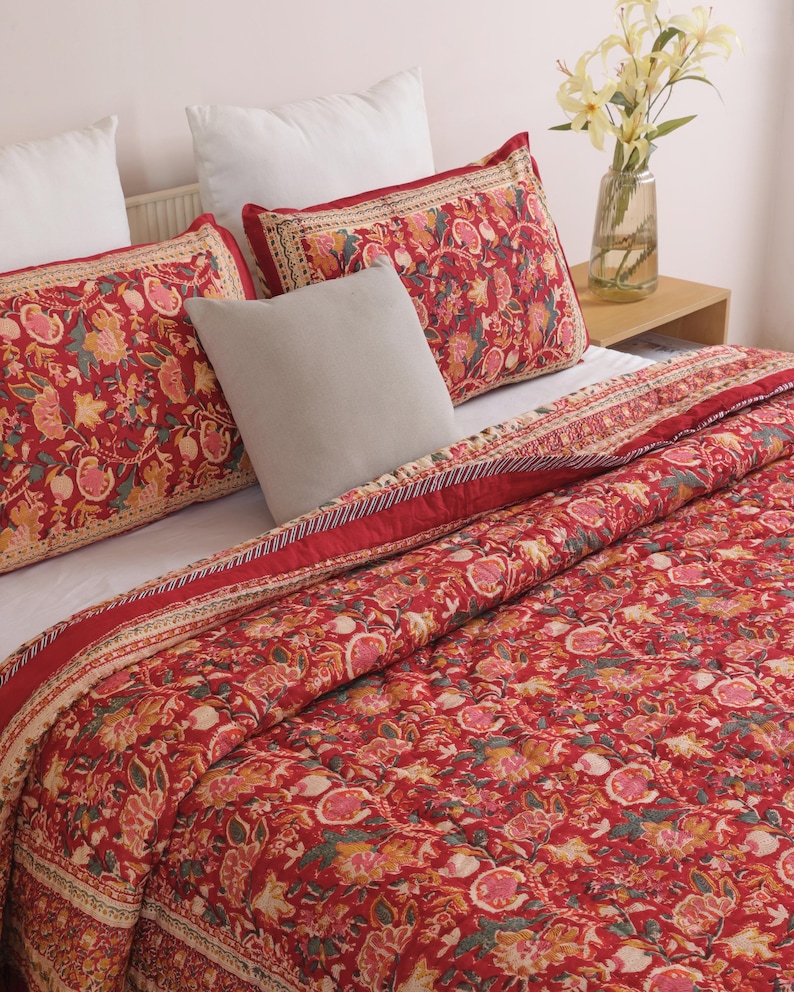 May include: A red quilted bedspread and two matching rectangular pillow shams featuring a dense floral and pomegranate pattern in shades of pink, orange, and green. A solid light gray square throw pillow sits between the two patterned shams.