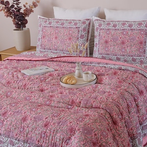 Peut inclure: Une courtepointe rose à motifs floraux et deux taies d'oreiller assorties sur un lit. La literie présente un motif répété de fleurs et de feuilles dans des tons de rose, de violet et de gris. Un plateau avec un croissant et une tasse de café est posé sur la courtepointe.