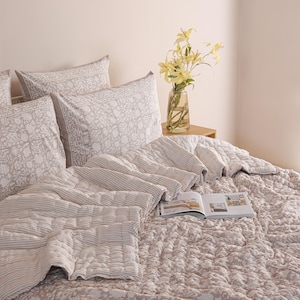 May include: A bed is made with a floral patterned comforter and matching pillowcases. A striped quilt and pillows are arranged on top. A vase of yellow flowers sits on a small wooden table. An open book rests on the bed.