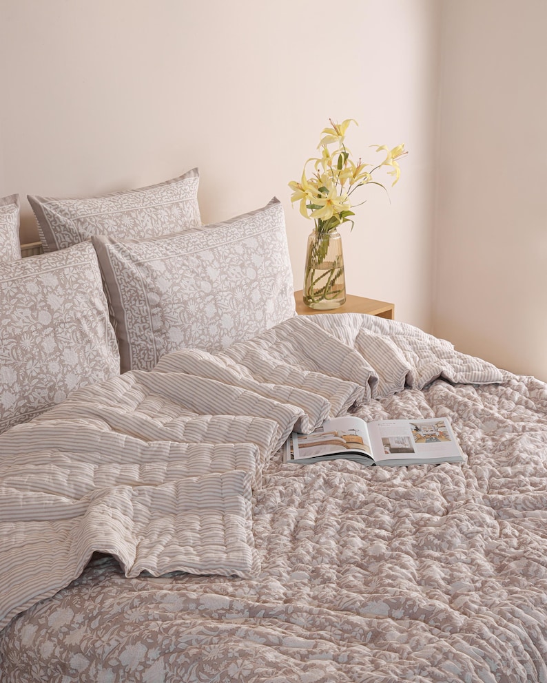 May include: A bed is made with a floral patterned comforter and matching pillowcases. A striped quilt and pillows are arranged on top. A vase of yellow flowers sits on a small wooden table. An open book rests on the bed.