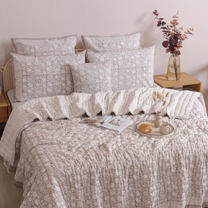 May include: A bed with a floral patterned comforter, pillows, and a decorative throw. The bedding is in shades of gray and white. A small wooden side table holds a vase of flowers and a tray with a croissant and coffee cup.