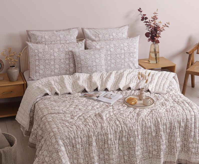 May include: A bed with a floral patterned comforter, pillows, and a decorative throw. The bedding is in shades of gray and white. A small wooden side table holds a vase of flowers and a tray with a croissant and coffee cup.