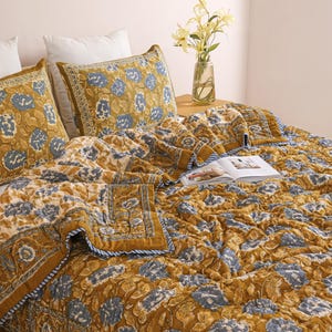 Może przedstawiać: Zestaw pościeli z kwiatowym wzorem w odcieniach musztardowej żółci i niebieskiego. Zestaw zawiera kołdrę i dwie poszewki na poduszki. Kołdra ma niebiesko-białą obwódkę w paski. Widoczny jest również wazon z kwiatami i otwarta książka.