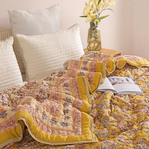 Puede incluir: Una cama con un edredón floral amarillo y fundas de almohada a juego. El edredón tiene un borde amarillo y un fondo crema con estampados florales y geométricos en tonos rosa, azul y verde. Un jarrón con flores amarillas está sobre una mesita de noche.