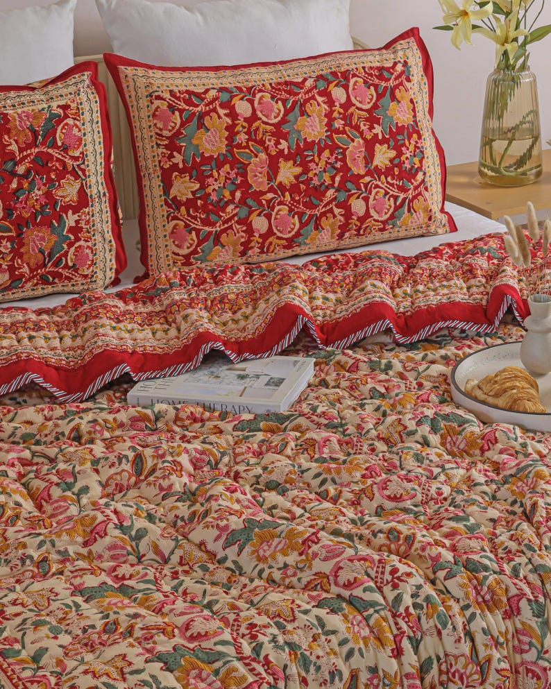 May include: A quilted bedspread and two matching rectangular pillow shams featuring a vibrant red, cream, green, and yellow floral block print pattern. The bedding set displays a detailed botanical motif with a decorative border on the pillows.