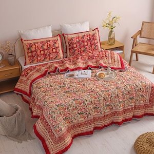 May include: A quilted bedspread and two matching pillow shams featuring a cream-colored base with a dense, multi-colored floral and vine pattern in shades of red, orange, green, and yellow, finished with a wide, solid red border trim.