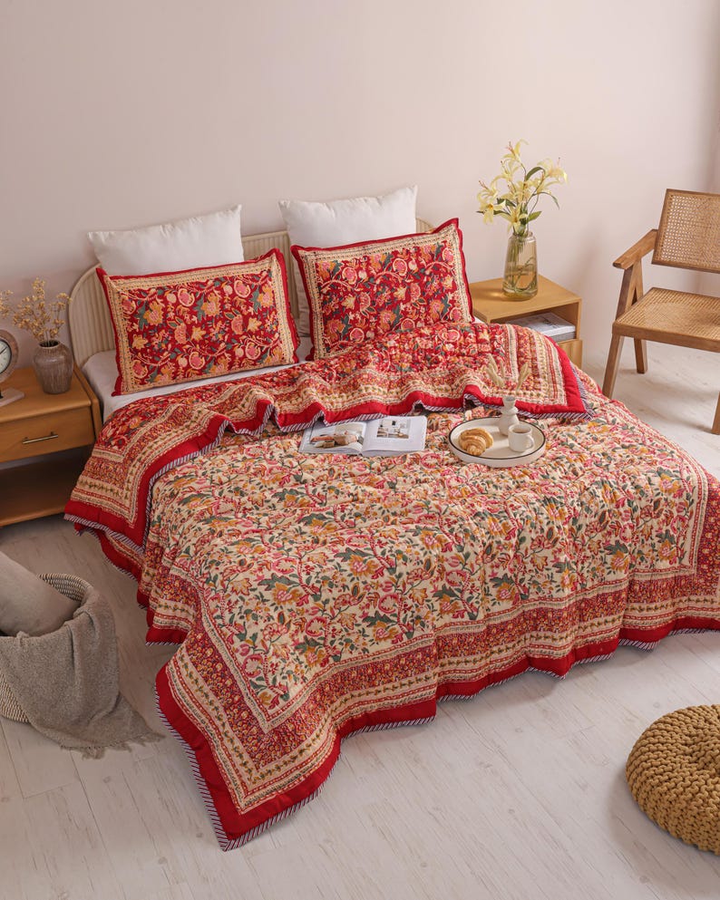 May include: A quilted bedspread and two matching pillow shams featuring a cream-colored base with a dense, multi-colored floral and vine pattern in shades of red, orange, green, and yellow, finished with a wide, solid red border trim.