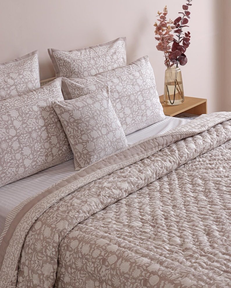 May include: A bed set featuring a floral patterned comforter, pillowcases, and pillows in shades of beige and white. The bedding is arranged on a bed with a striped sheet. A vase with decorative branches sits on a wooden nightstand.