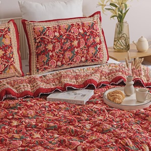 May include: A red quilted bedding set featuring a repeating floral pattern in shades of pink, green, and cream. The set includes a large rectangular pillow sham and a matching quilt. A book titled Home Therapy is placed on the textured fabric surface.