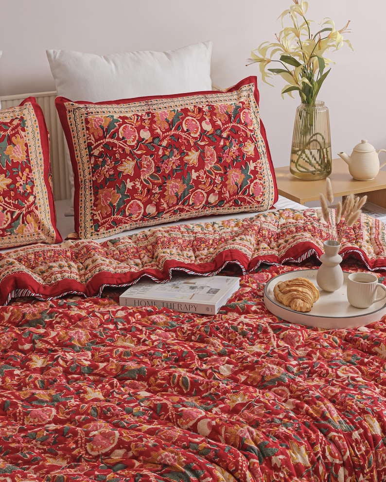 May include: A red quilted bedding set featuring a repeating floral pattern in shades of pink, green, and cream. The set includes a large rectangular pillow sham and a matching quilt. A book titled Home Therapy is placed on the textured fabric surface.
