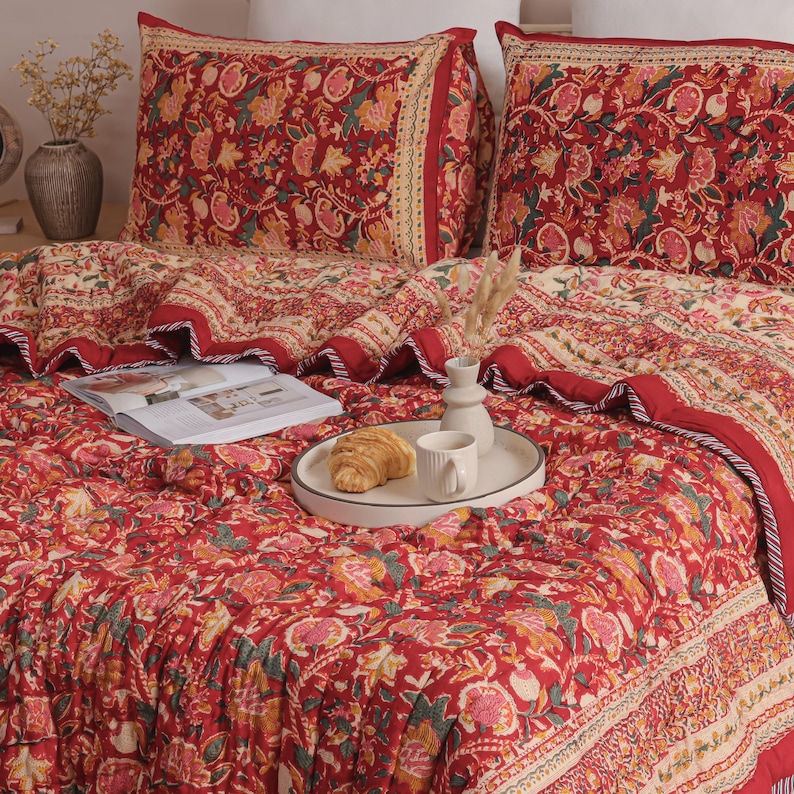 May include: A quilted bedding set featuring a vibrant red base with a repeating floral pattern in shades of yellow, pink, and green. The set includes two matching rectangular pillow shams and a large quilt, all finished with a decorative border.