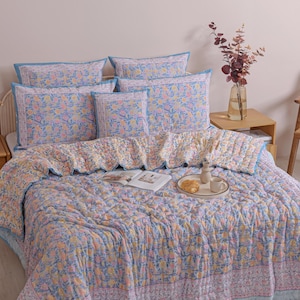 May include: A bed is covered with a floral patterned quilt and matching pillows. The quilt and pillows have a light blue background with a repeating pattern of orange and yellow flowers. The bed is made up with a light blue bed skirt.