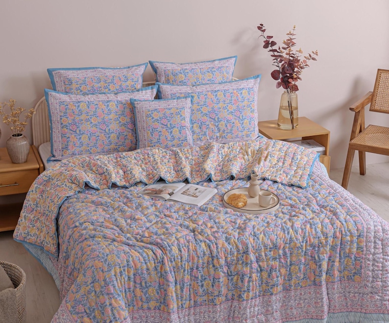 May include: A bed is covered with a floral patterned quilt and matching pillows. The quilt and pillows have a light blue background with a repeating pattern of orange and yellow flowers. The bed is made up with a light blue bed skirt.